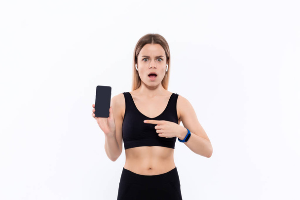 Surpreendido jovem mulher loira desportiva em um sportswear preto com botões de ouvido sem fio mostrando tela de um smartphone com um dedo em pé sobre fundo branco
 - Foto, Imagem