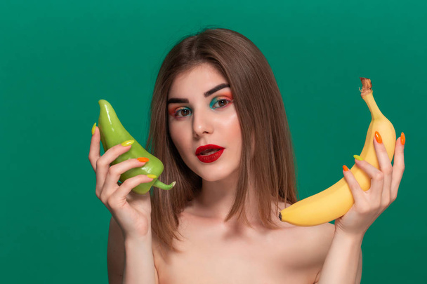 Крупним планом портрет красивої молодої жінки з яскравим кольоровим макіяжем, що тримає зелений перець і жовтий банан, що стоїть ізольовано на зеленому фоні
 - Фото, зображення