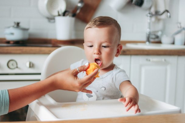 Um bebé encantador a comer pêssego na cozinha. Mãe alimenta criança
 - Foto, Imagem