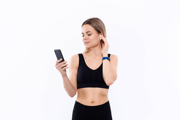 Excited young sporty blond woman in a black sportswear with wireless ear buds looking at the screen of a smartphone standing isolated over white background. - Photo, image