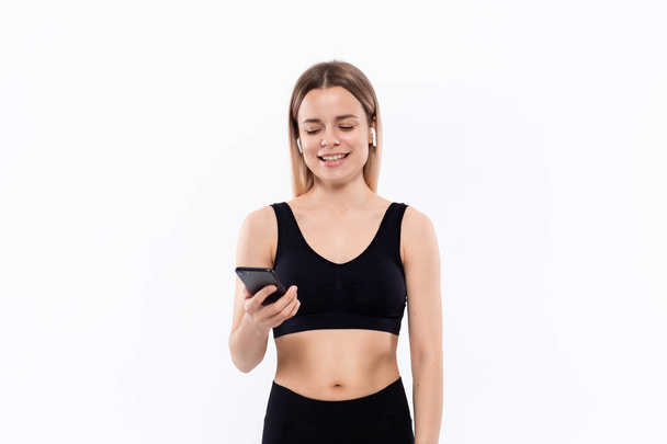 Excited young sporty blond woman in a black sportswear with wireless ear buds looking at the screen of a smartphone standing isolated over white background. - Fotoğraf, Görsel