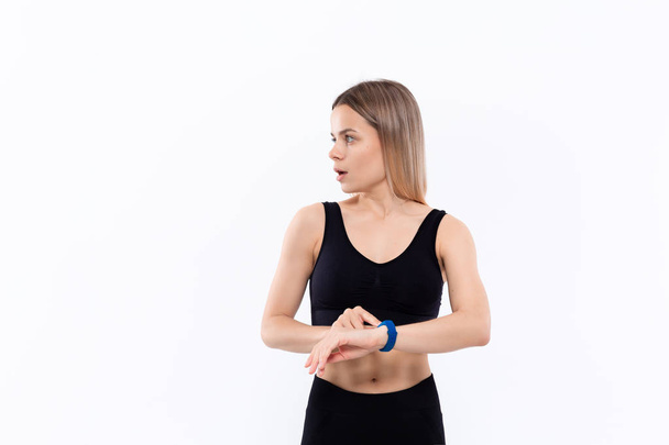 Mladá sportovně plavovlasá žena v černém sportovním přípoji překvapeně překvapila pulz s inteligentními hodinkami, když se rozpracovala nad bílým pozadím. - Fotografie, Obrázek