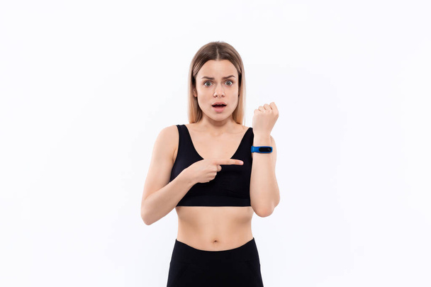 Giovane donna bionda sportiva in uno sportswear nero sorpreso controllare il polso con orologi intelligenti dopo l'allenamento in piedi su sfondo bianco
 - Foto, immagini