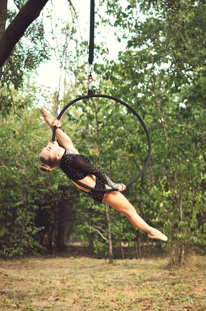 Mladá akrobatka v černém obleku s rhinestony cvičením na vzdušném akrobacickém kruhu, který visí na stromě v letním parku. - Fotografie, Obrázek