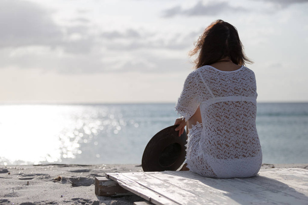 Mujer joven por detrás sentada junto al mar mira al horizonte al amanecer en el viento, vestida con un vestido de encaje blanco y ropa interior blanca y pelo largo
 - Foto, Imagen