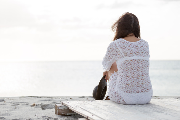 Mujer joven por detrás sentada junto al mar mira al horizonte al amanecer en el viento, vestida con un vestido de encaje blanco y ropa interior blanca y pelo largo
 - Foto, imagen