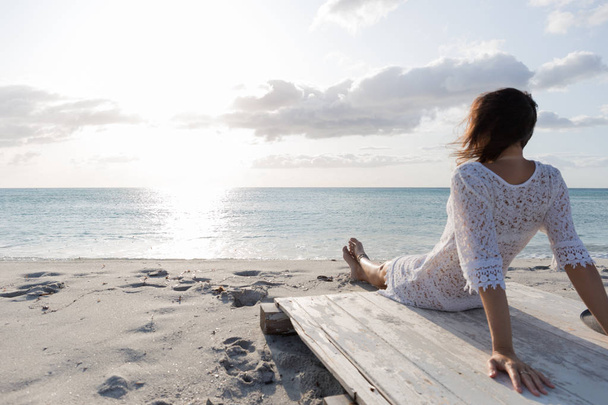 Jonge vrouw van achteren zittend aan de zee kijkt naar de horizon bij zonsopgang in de wind, gekleed in een witte kanten jurk en wit ondergoed en lang haar - Foto, afbeelding