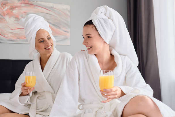 Two young beautiful women in white bathrobes and towels on head happily talking together holding glasses with orange juice in hands at modern home - Zdjęcie, obraz