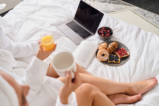 Close up photo of women holding glass of juice and cup of coffee with laptop, book and beautiful breakfast in bed in modern hotel  - Foto, afbeelding