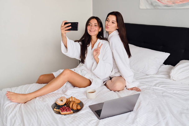 Twee aantrekkelijke vrouwen in witte badjassen die gelukkig Foto's samen nemen op mobiele telefoon met heerlijk gebak en koffie. Mooie meiden ontbijten in bed met laptop in modern hotel - Foto, afbeelding