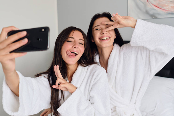 Молодые симпатичные улыбающиеся женщины в белых халатах с радостью показывают два пальца жест делая классные фотографии вместе на мобильном телефоне в уютном доме
  - Фото, изображение