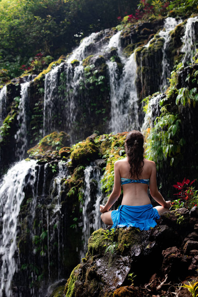Молодая белая женщина медитирует, практикует йогу у водопада. Вид сзади. Водопад Banyu Wana Amertha Wanagiri, Бали, Индонезия
.  - Фото, изображение