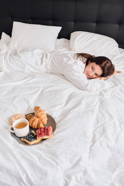 Mladá krásná žena v bílém županu leží ve velké posteli s lahodnými pečivo a šálkem čaje na snídani poblíž moderního útulného hotelu - Fotografie, Obrázek