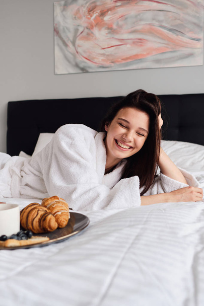 白いバスローブを着た若い陽気な女性は、モダンな居心地の良いホテルの近くで朝食においしいペストリーでベッドに横たわって笑っています - 写真・画像