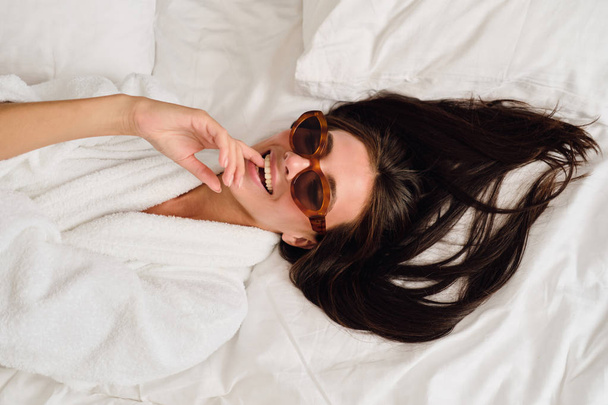 Молодая красивая улыбающаяся женщина с темными волосами в белом халате и солнечных очках мечтательно кусает палец, лежащий на кровати в современном уютном отеле
 - Фото, изображение