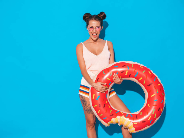 Young beautiful sexy smiling hipster woman in sunglasses.Girl in summer clothes with donut lilo inflatable mattress.Positive female going crazy.Funny model isolated on blue - Photo, image