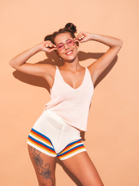 Portrait of young beautiful sexy woman with two horns hairstyle. Trendy girl in casual summer hipster knitted cardigan topic ,shorts in sunglasses. Hot model isolated beige wall. - Fotó, kép