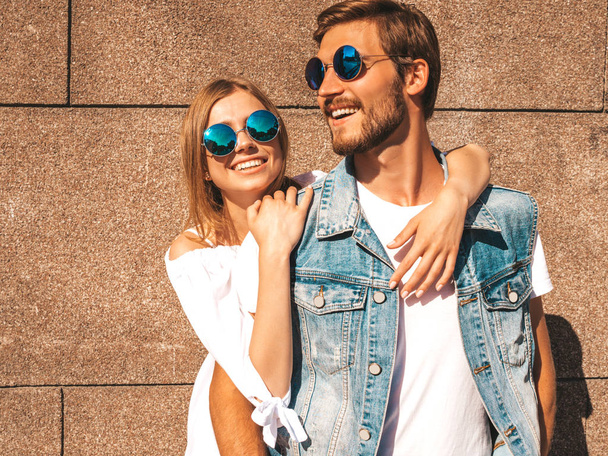 Smiling beautiful girl and her handsome boyfriend. Woman in casual summer dress.Man in jeans clothes.Happy cheerful family.Female having fun on the street near wall.Hugging couple in sunglasses - Fotó, kép
