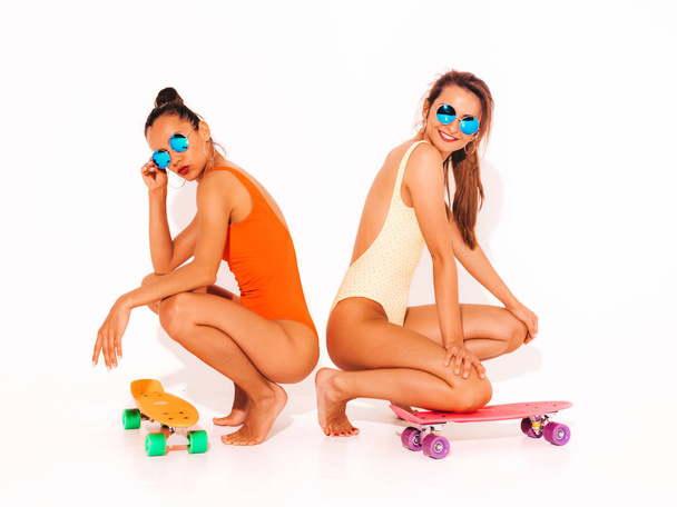 Two beautiful sexy smiling hipster women in summer colorful swimwear bathing suits.Trendy Girls in sunglasses. Positive models sitting on the floor with colorful penny skateboards.Isolated on white - Fotografie, Obrázek