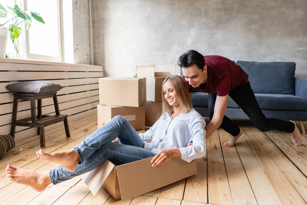 Happy couple having fun and riding in cardboard boxes at new home. Young couple moving to a new apartment together. Relocation concept - Fotoğraf, Görsel