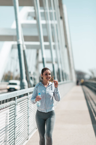 Beautiful young and fit woman in good shape running and jogging alone on city bridge street. She wears sporty earbuds and listens to music. - Foto, Imagen