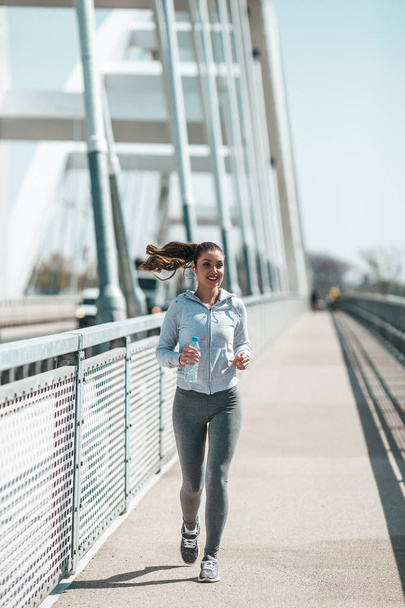 Beautiful young and fit woman in good shape running and jogging alone on city bridge street. She wears sporty earbuds and listens to music. - Foto, Bild