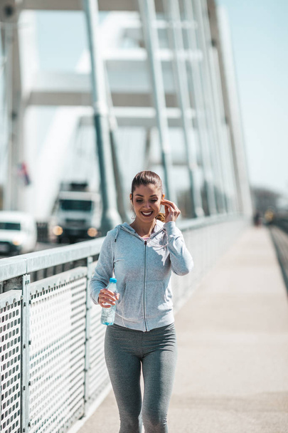 Beautiful young and fit woman in good shape running and jogging alone on city bridge street. She wears sporty earbuds and listens to music. - Photo, Image