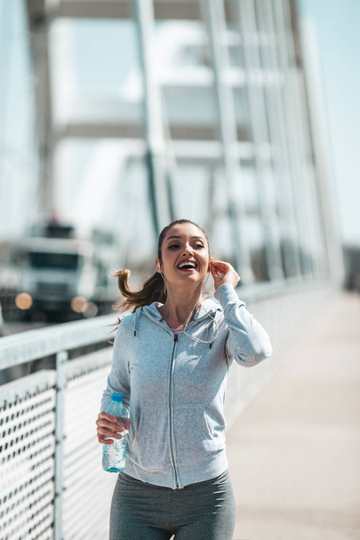 Beautiful young and fit woman in good shape running and jogging alone on city bridge street. She wears sporty earbuds and listens to music. - Photo, image