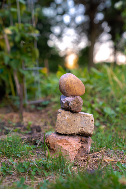 Een stapel of stapel kleine rotsen met Earth Tone kleuren gelaagd op elkaar en zittend in het gras in de buurt van een tuin met wazig bokeh achtergrond met zon licht schijnt door de bomen voorbij. - Foto, afbeelding