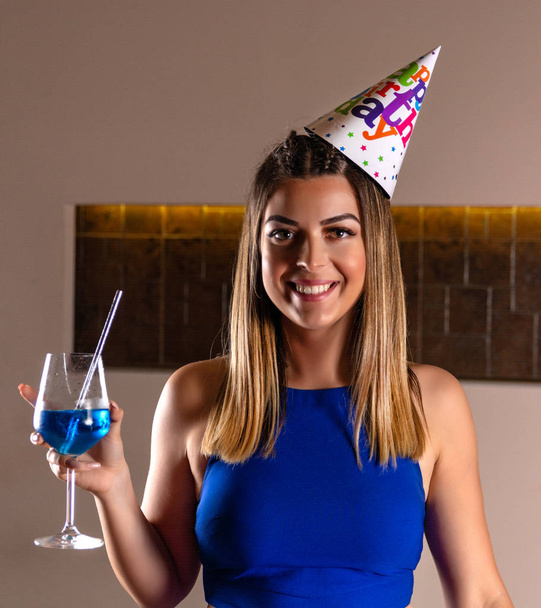 Happy girl with birthday cap on head and blue lagoon cocktail in hand at a night club. Birthday party concept. Close up, selective focus - Photo, Image