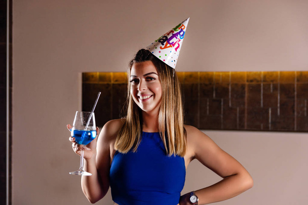ナイトクラブで手に頭と青いラグーンカクテルの誕生日キャップを持つ幸せなかわいい女の子。誕生日パーティーのコンセプト。クローズアップ、選択的フォーカス - 写真・画像
