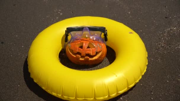 pumpkin lantern on the beach in a diving mask and a yellow rubber ring on the sea background - Footage, Video