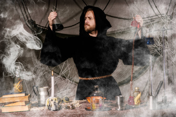 Ο μεσαιωνικός αλχημιστής κάνει μαγική τελετουργία στο τραπέζι στο εργαστήριο καπνού του. - Φωτογραφία, εικόνα