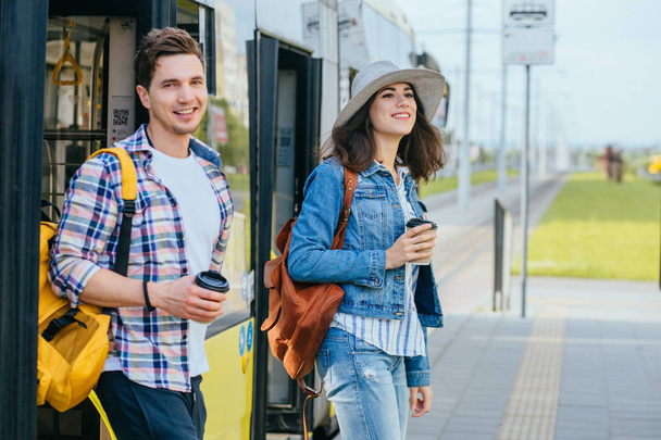 Lifestyle photo of young woman and man come out while opening door to tram. Female traveler In denim jacket with backpack smiling and talking with handsome guy on tram stop. - Foto, afbeelding