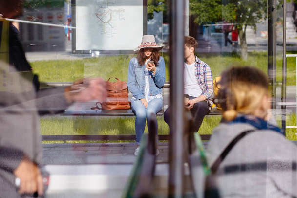 Photo à travers la fenêtre du tram : jeune couple voyageur assis sur la station de tram ou s'arrêter en plein air avec des smartphones
. - Photo, image