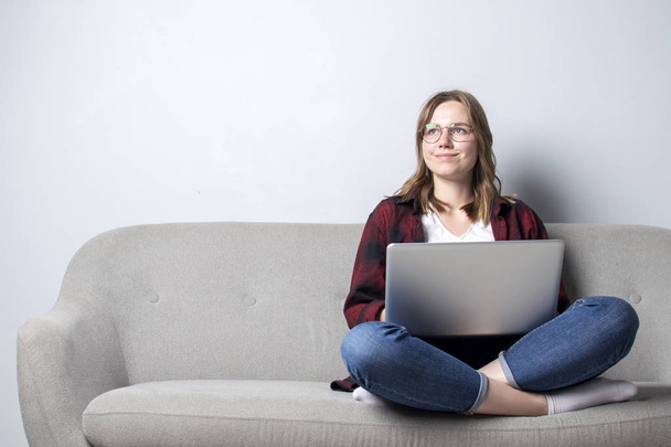 Młoda dziewczyna z laptopem siedzi na kanapie i picie kawy, Kobieta za pomocą komputera przed białą pustą ścianę, ona Freelancing i Dreaming, Kopiuj przestrzeń - Zdjęcie, obraz