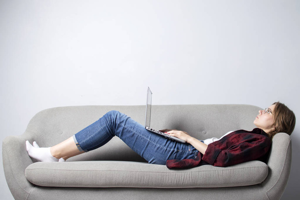 young girl with a laptop lying on the couch and relax, a woman using a computer against a white blank wall, she freelancing and printing text, copy space - Photo, image