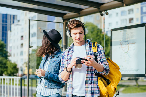 Porträt eines lächelnden Teenagers mit schwarzem Hut, Jeansjacke, die mit Freund an der Bushaltestelle hängt und Mobiltelefone benutzt. Freiheit, Reisekonzept. - Foto, Bild
