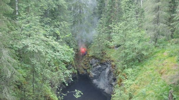 Вид с воздуха на человека, стоящего на краю глубокого ущелья с красной сигнальной вспышкой в руке в лесу возле высоких старых деревьев и кустарников. Запись. SOS сигналы в лесу
 - Фото, изображение