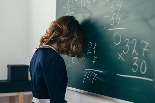 Sad girl pupil trying to solve an example. Schoolgirl stands with her forehead on the blackboard - Φωτογραφία, εικόνα