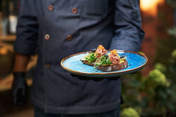 delicious raw beef tartare on bruschette, the dish is held in hand by a cook in a blue uniform. - Foto, afbeelding