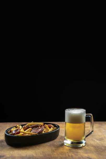foam in a glass with beer and fried sausages with potatoes on a wooden background.Oktoberfest. - Foto, Imagen