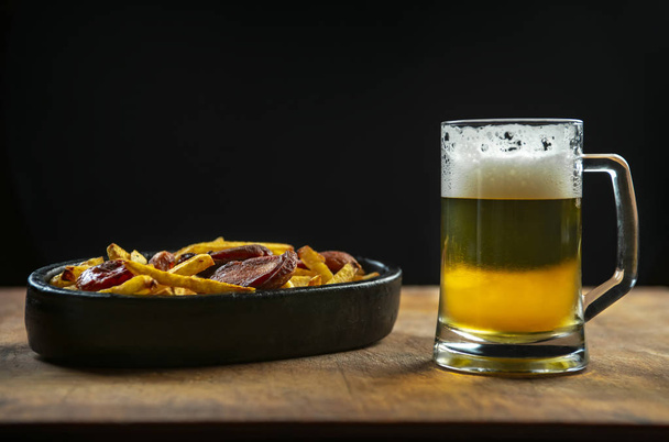 пена в стакане с пивом и жареные колбасы с картофелем на деревянном фоне.. - Фото, изображение