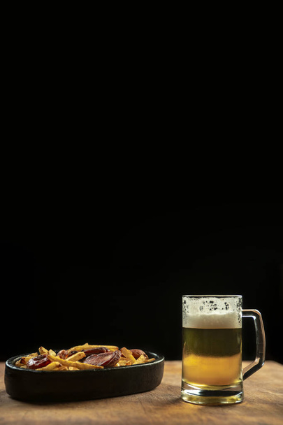 foam in a glass with beer and fried sausages with potatoes on a wooden background.Oktoberfest. - Foto, Bild