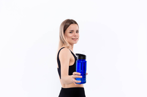 junge sportliche blonde Frau in schwarzer Sportbekleidung mit smarten Uhren hält Flasche mit Wasser nach dem Training vor weißem Hintergrund. - Foto, Bild