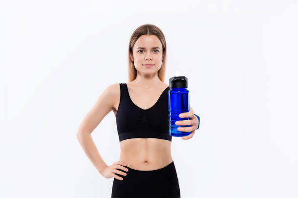 Jovem mulher loira desportiva em um sportswear preto com relógios inteligentes mantém garrafa com água após o treino em pé sobre fundo branco
. - Foto, Imagem