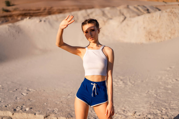 A beautiful girl in a sexy white top and short shorts stands in the desert and covers her eyes with the hand from the sun. A hand shadow falls on her face. - Foto, imagen