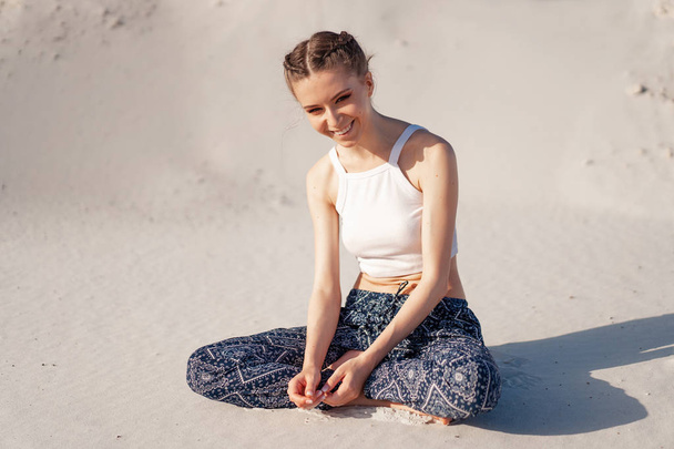 A beautiful young Caucasian girl in a white top and wide pants sits in a lotus position on the beach on the sand.  - Zdjęcie, obraz