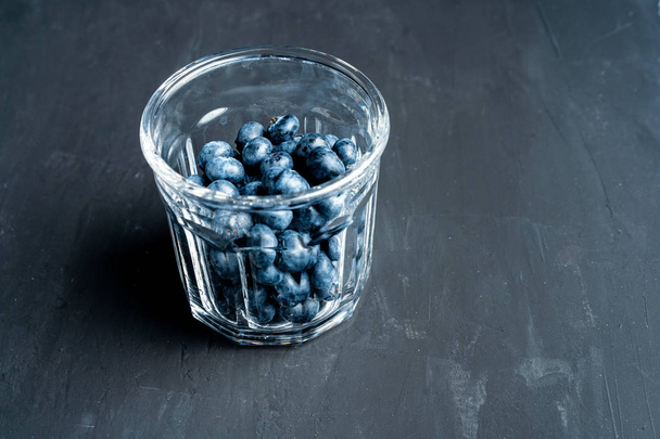 Beautiful tasty raw blueberry with a transparent glass cup on a dark black concrete background. - Valokuva, kuva