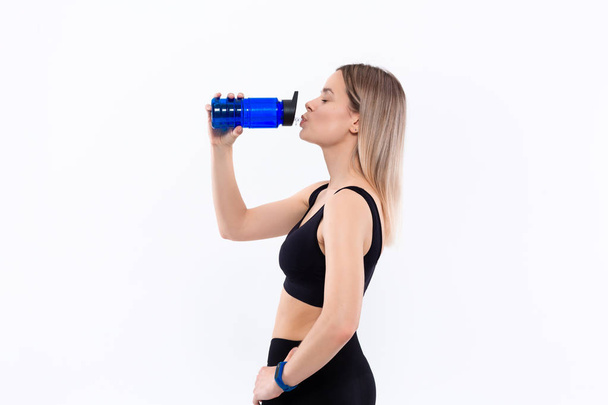 junge sportliche blonde Frau in schwarzer Sportbekleidung mit smarten Uhren trinkt nach dem Training vor weißem Hintergrund stehend Wasser. - Foto, Bild